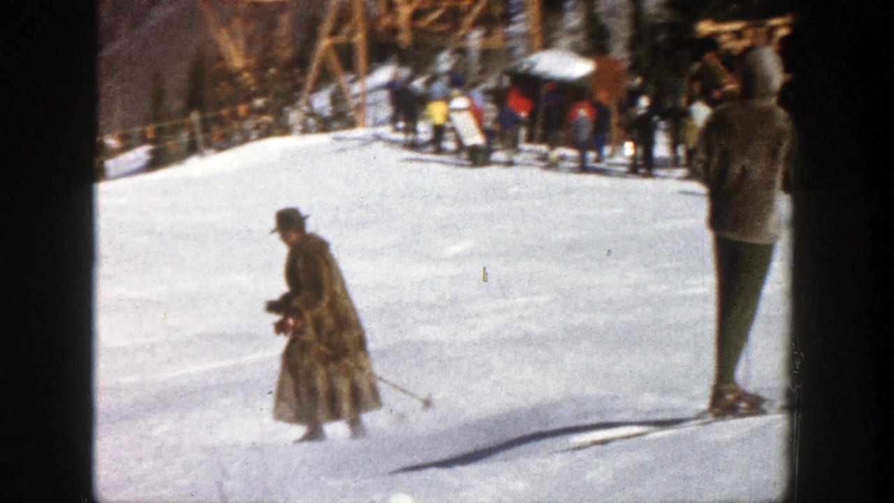 Skier in a long coat moves across a snowy slope with a standing skier at the right edge.