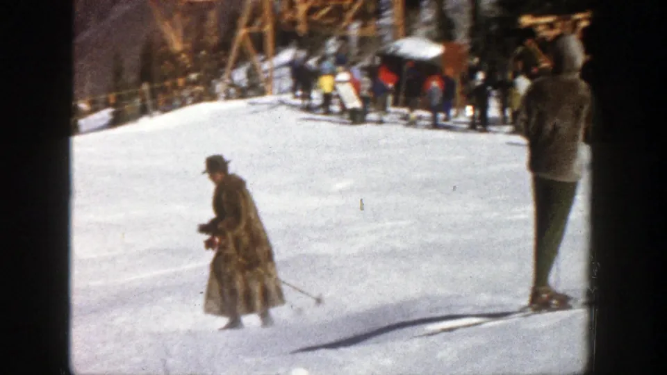 1961 Aspen frame of a long-coated skier using poles on the snow, with other skiers and a rustic structure in the background.