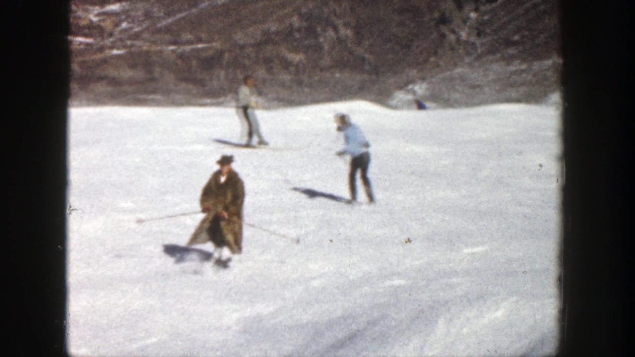 1961 Aspen, Colorado home-movie frame showing a man in a brown fur coat skiing backward while other adults ski behind him.
