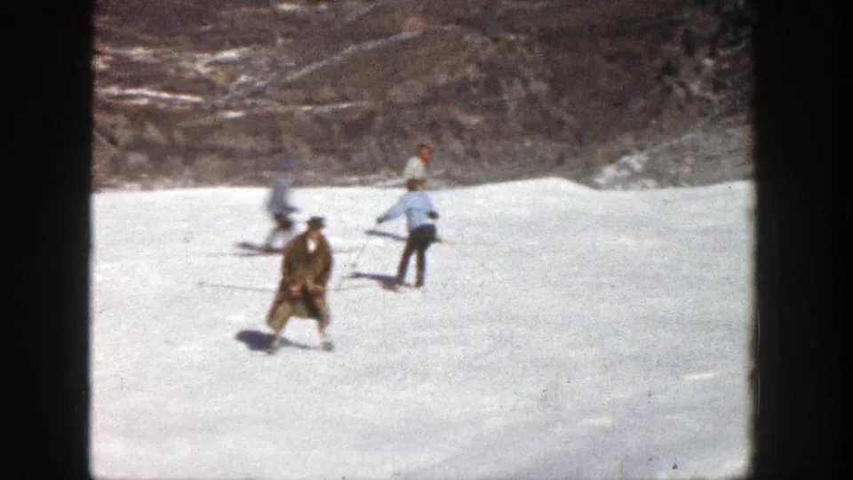 Aspen ski-slope frame from 1961 showing the brown-coated skier facing the camera area while other adults pass behind him.
