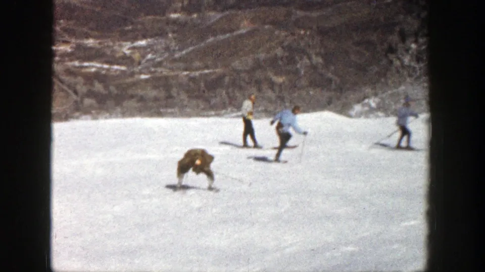 1961 Aspen home-movie frame showing the foreground skier crouched low on the slope with other skiers passing behind him.