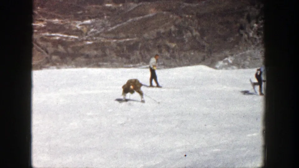 Aspen, Colorado 1961 frame with the brown-coated skier bent forward on skis and another adult skier upright in the background.