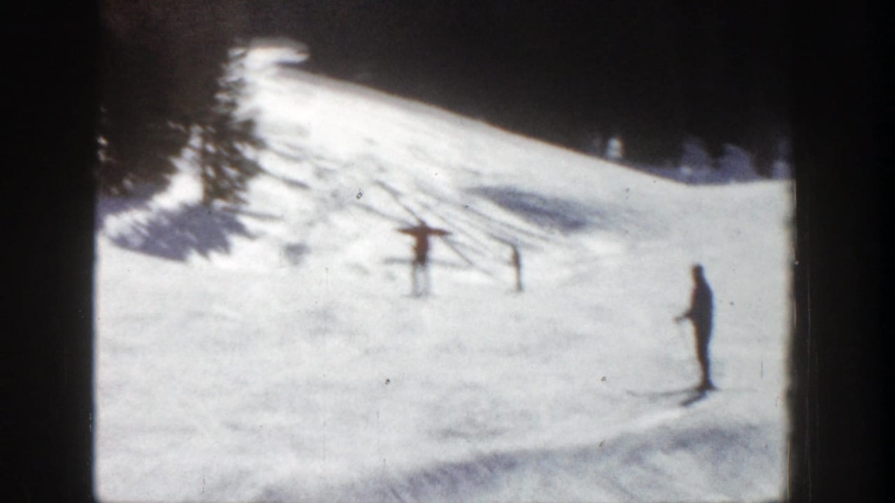 Distant skier on a sunlit Aspen ski slope with simple fencing, trees, and snow-covered ground visible.