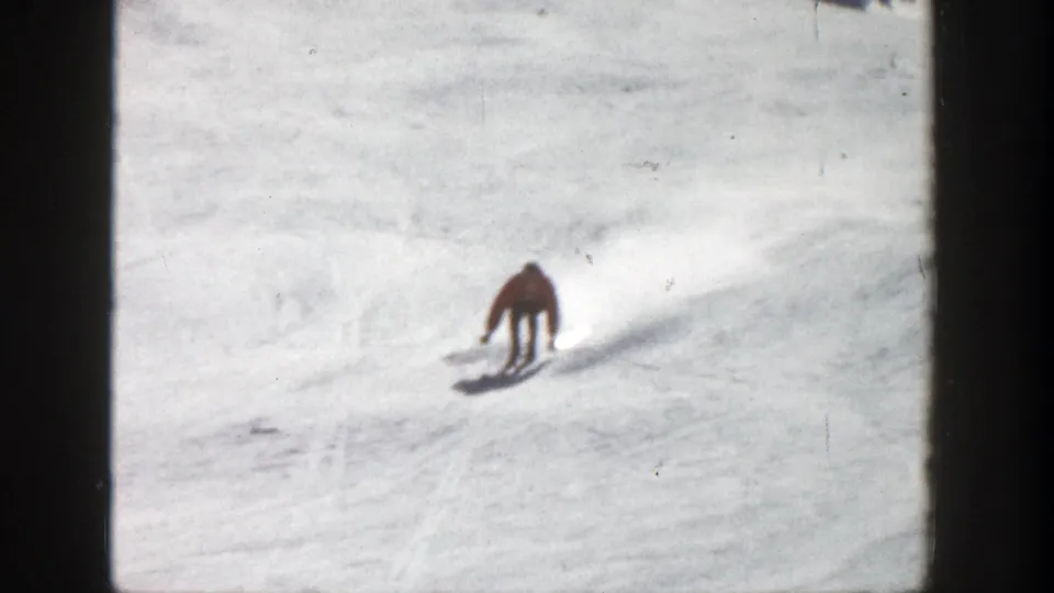 Skier in a red jacket descends a snowy Aspen slope in soft 8mm color with long shadows visible.