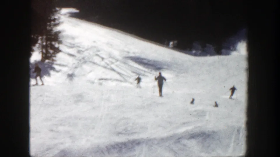 Distant skiers cross a bright Aspen ski slope, with dark trees at the ridge and soft 8mm home-movie texture.