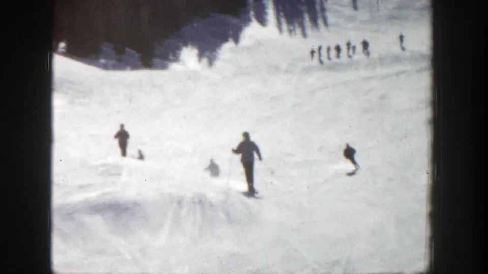 A foreground skier moves down the Aspen slope while other small figures gather or ski near the upper snowfield in 1961 footage.