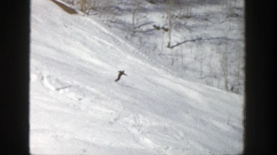 A small figure skis across the lower part of the snowy Aspen run, with tracks and tree shadows visible in the 8mm frame.