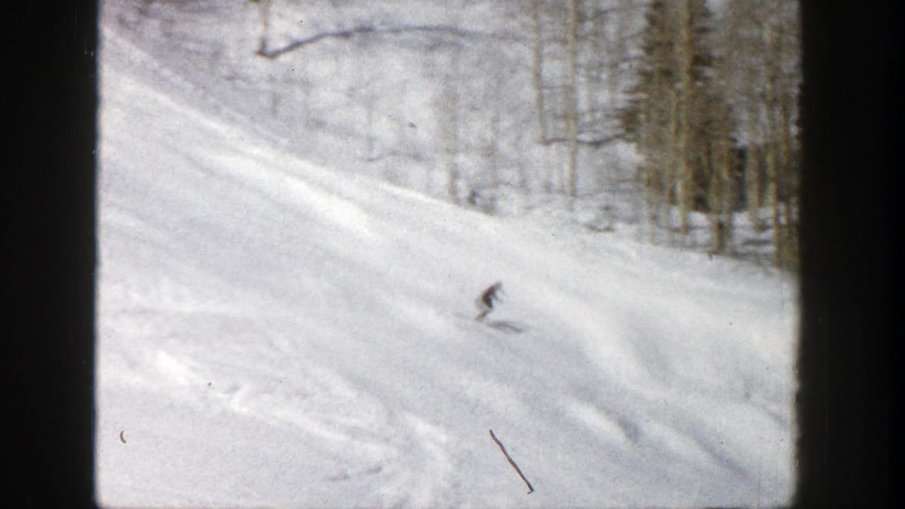 A distant skier moves downhill on a snowy Aspen slope in 1961, framed by bare trees and 8mm film grain.