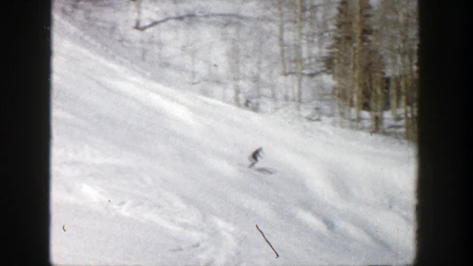 A distant skier moves downhill on a snowy Aspen slope in 1961, framed by bare trees and 8mm film grain.
