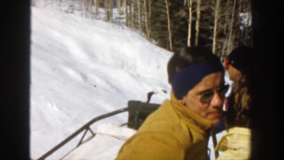 A man wearing sunglasses, a blue headband, and a yellow sweater sits on a ski lift in Aspen, with snowy trees behind him.