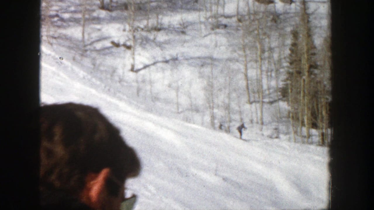 Partial spectator head in foreground with snowy ski slope and distant skier beyond.