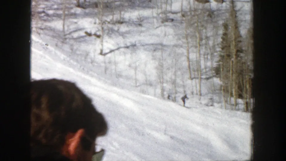 A partial spectator is seen in front of a snowy Aspen slope as a distant skier continues downhill.
