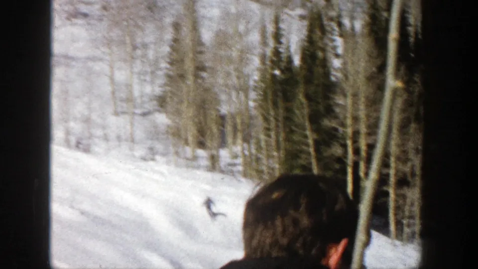 A distant skier crosses a snowy Aspen hillside while a partial spectator appears at the bottom edge of the frame.