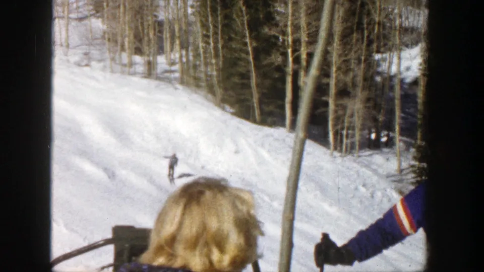 A blond spectator looks toward an Aspen ski run as a distant skier descends near trees and another figure appears at the edge.