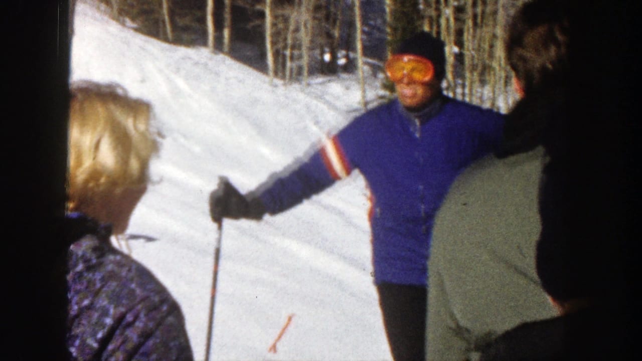 1961 Aspen frame of a skier in a blue jacket and orange goggles gesturing with poles beside other skiers on a snowy run.
