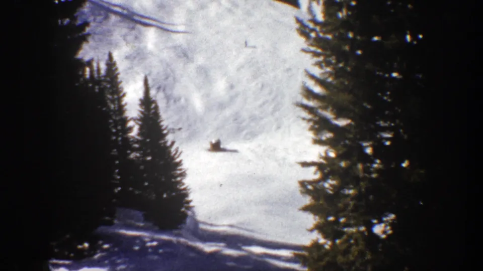 1961 Aspen home-movie frame showing a distant skier crossing a bright snowy slope between evergreen trees.