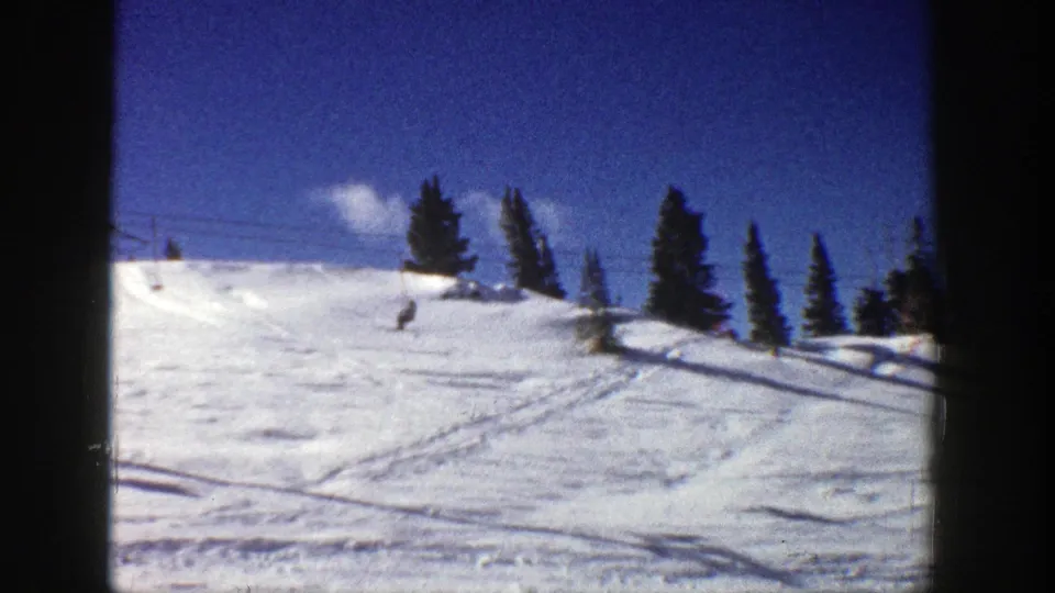 Wide 1961 Aspen winter frame with blue sky, evergreen trees, ski tracks, and distant people skiing downhill.