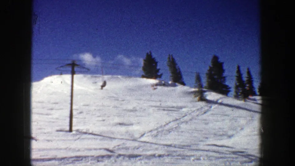 The 8mm frame shows a chairlift tower on a snowy Aspen slope, with a rider suspended against the hillside and trees beyond.