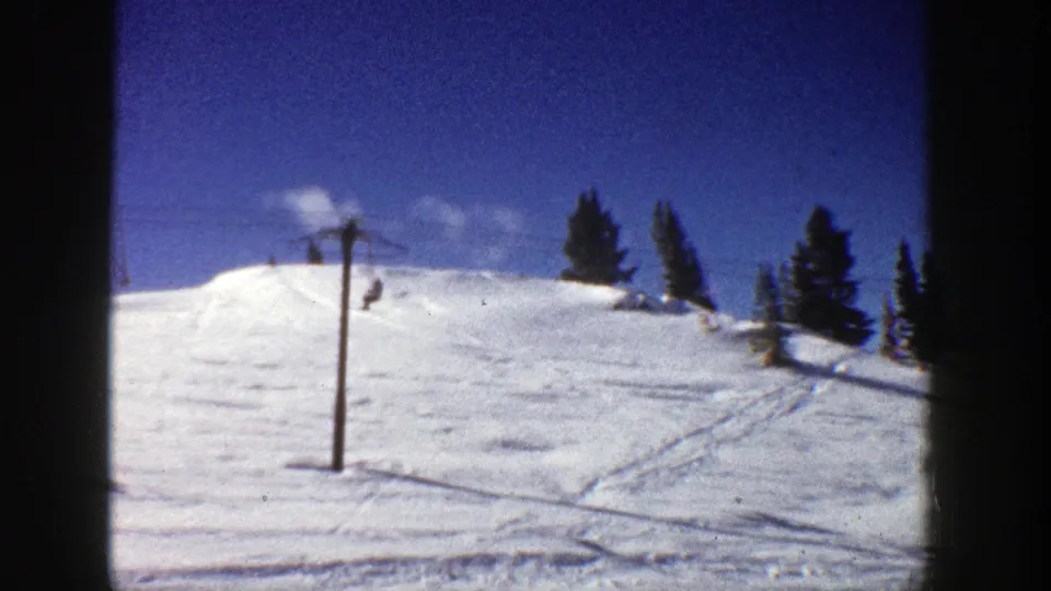 Distant chairlift riders move over the Aspen ski slope, with evergreen trees on the ridge and intersecting tracks in the snow.
