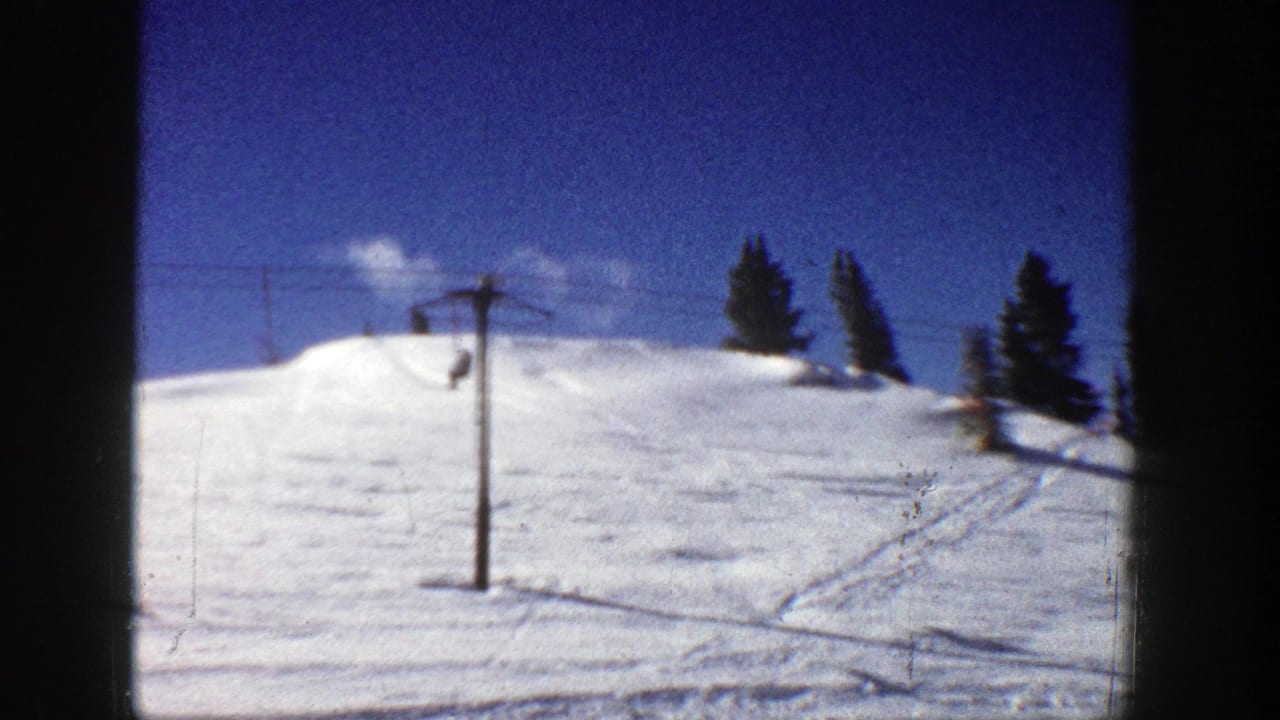 1961 Aspen ski slope with snow, a lift pole, evergreen trees, and clear blue sky in 8mm home-movie footage.