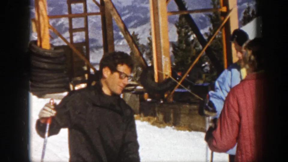 1961 Aspen home-movie frame of a man with glasses holding ski poles beside skiers at a snowy lift area.