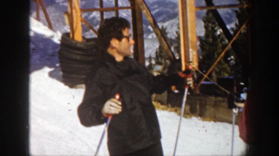 The man in dark winter clothing appears to turn while holding ski poles on a 1961 Aspen ski slope with trees behind him.