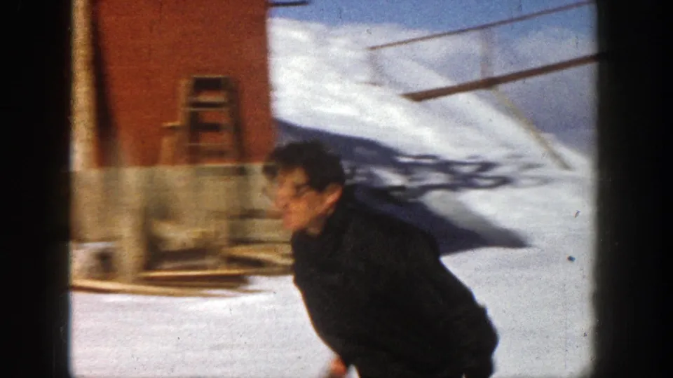 The skier bends forward as he starts downhill on the Aspen snow, with a red-brown wall and slope shadows visible behind him.