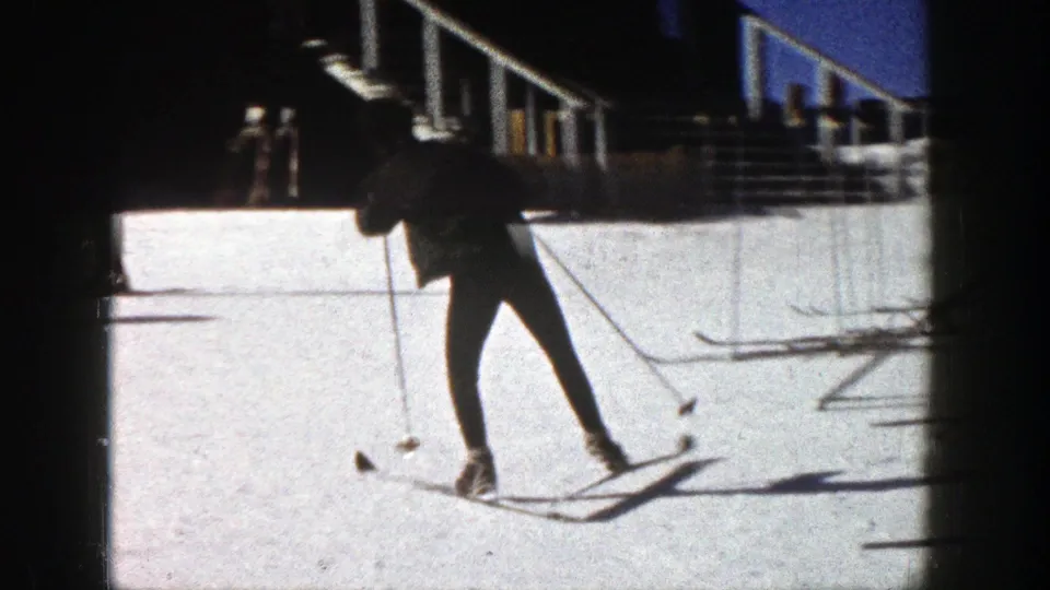 A skier in dark clothing practices with poles on a sunlit Aspen slope, with railings and a building edge in the background.