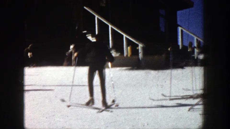 The skier continues across the Aspen snow, framed from farther back with poles, rails, and heavy 8mm edge shadowing visible.