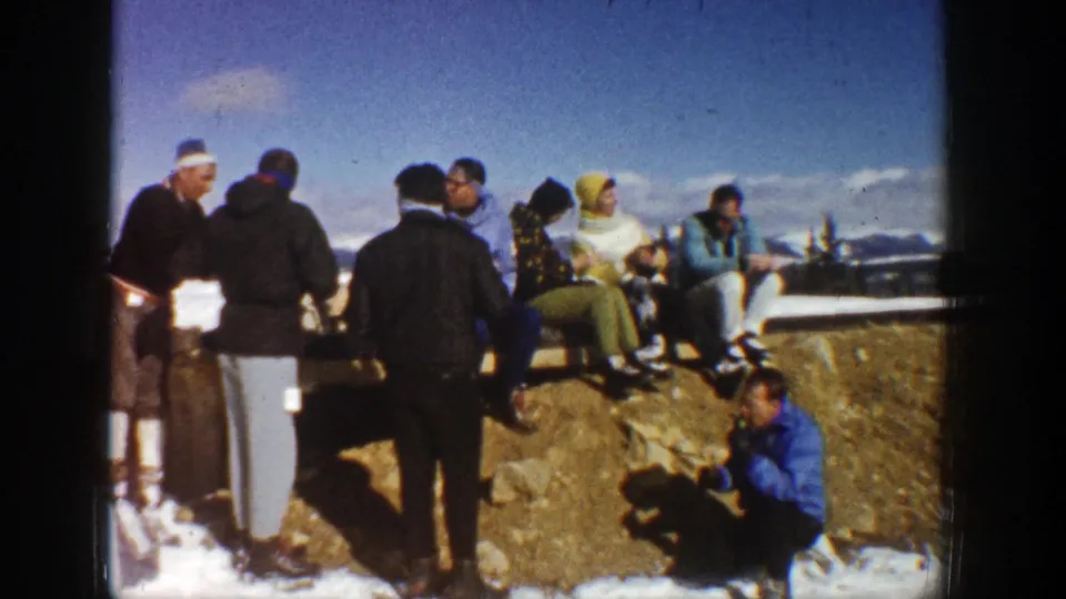 Bundled adults gather and sit on a rocky snowy overlook in Aspen, with blue sky and distant mountains behind them.