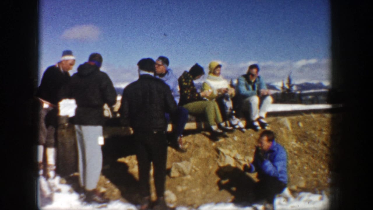 1961 Aspen home-movie frame showing adults in coats and hats resting and talking near a snowy rock ledge.