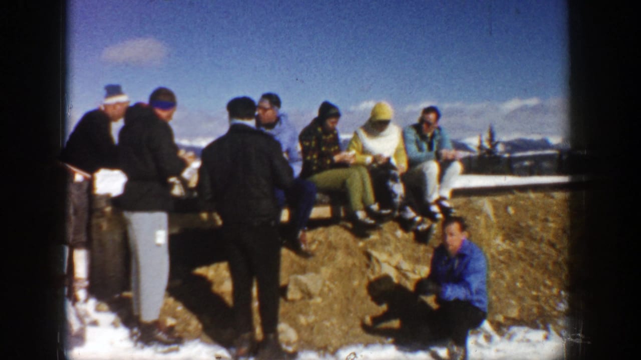 Group of adults sits and stands by a rocky ledge with snow and distant peaks visible.