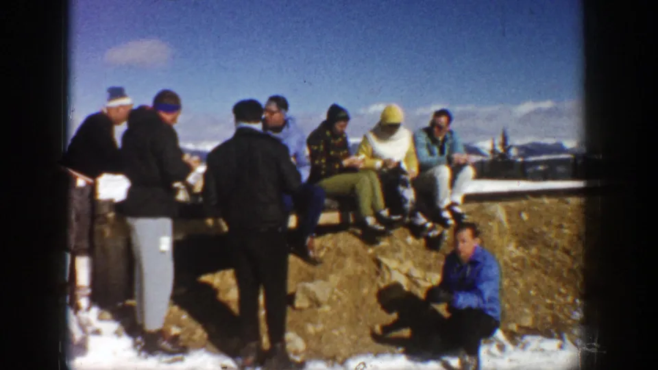 Adults in winter clothing gather on a rocky overlook in Aspen, Colorado, with snow patches and distant mountains visible.