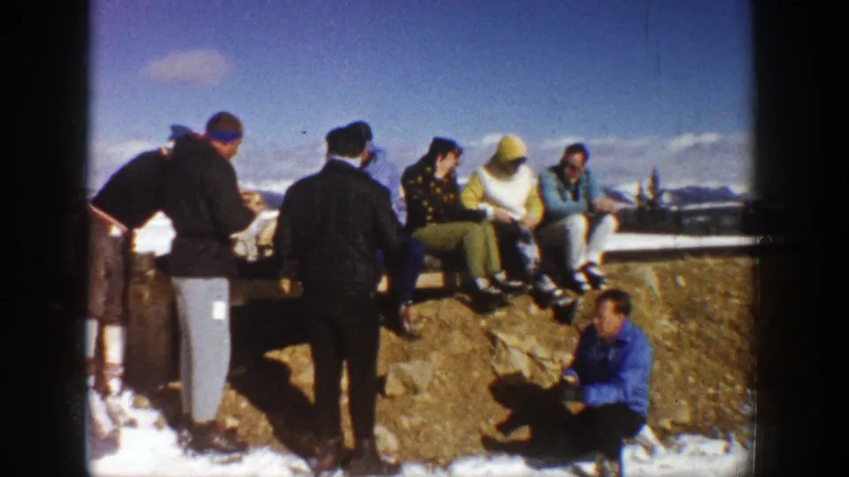 1961 Aspen frame showing adults gathered at a snow-dusted rock, including seated figures on the ledge and one person crouched below.