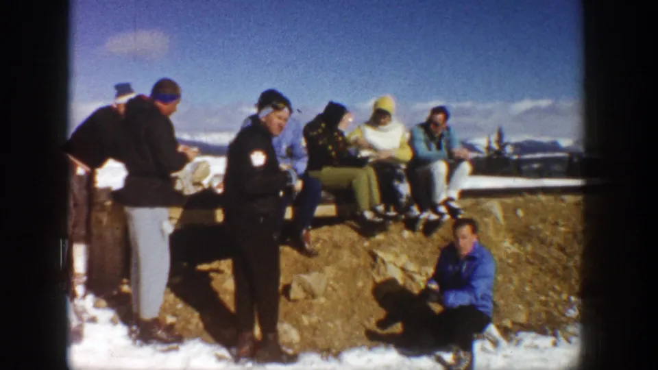 Adults at an Aspen mountain stop sit and stand near a snow-covered rock ledge, with distant peaks behind them.