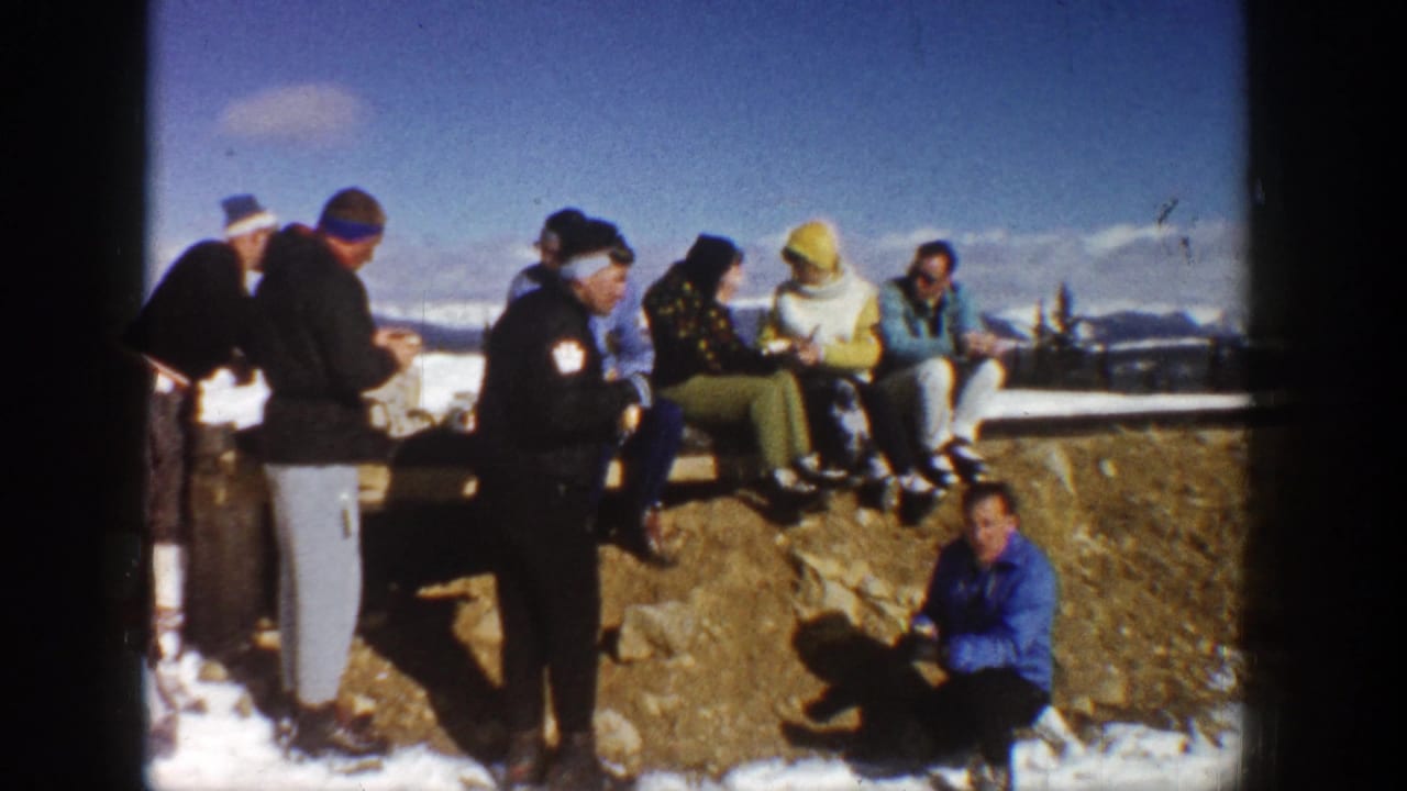 Adults in winter clothing gather by a snowy stone overlook with mountains in the distance.