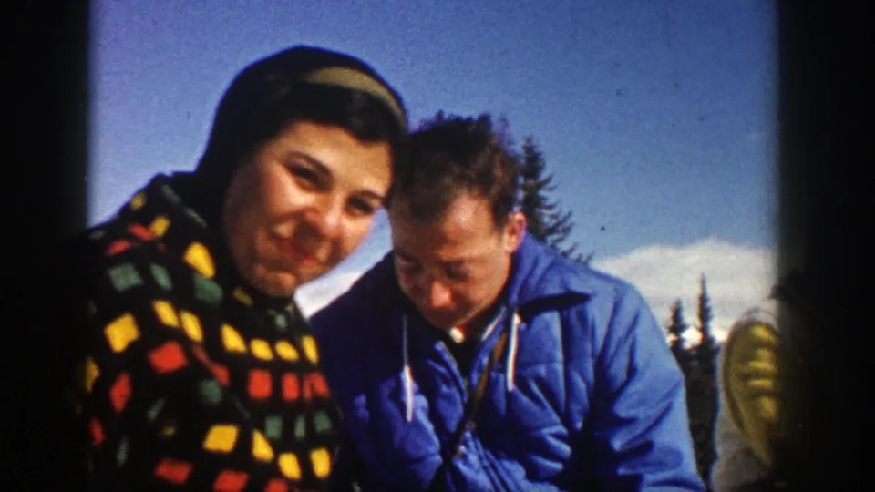 A close 1961 Aspen home-movie frame shows a woman in a bright patterned hooded coat beside a man in a blue winter jacket.