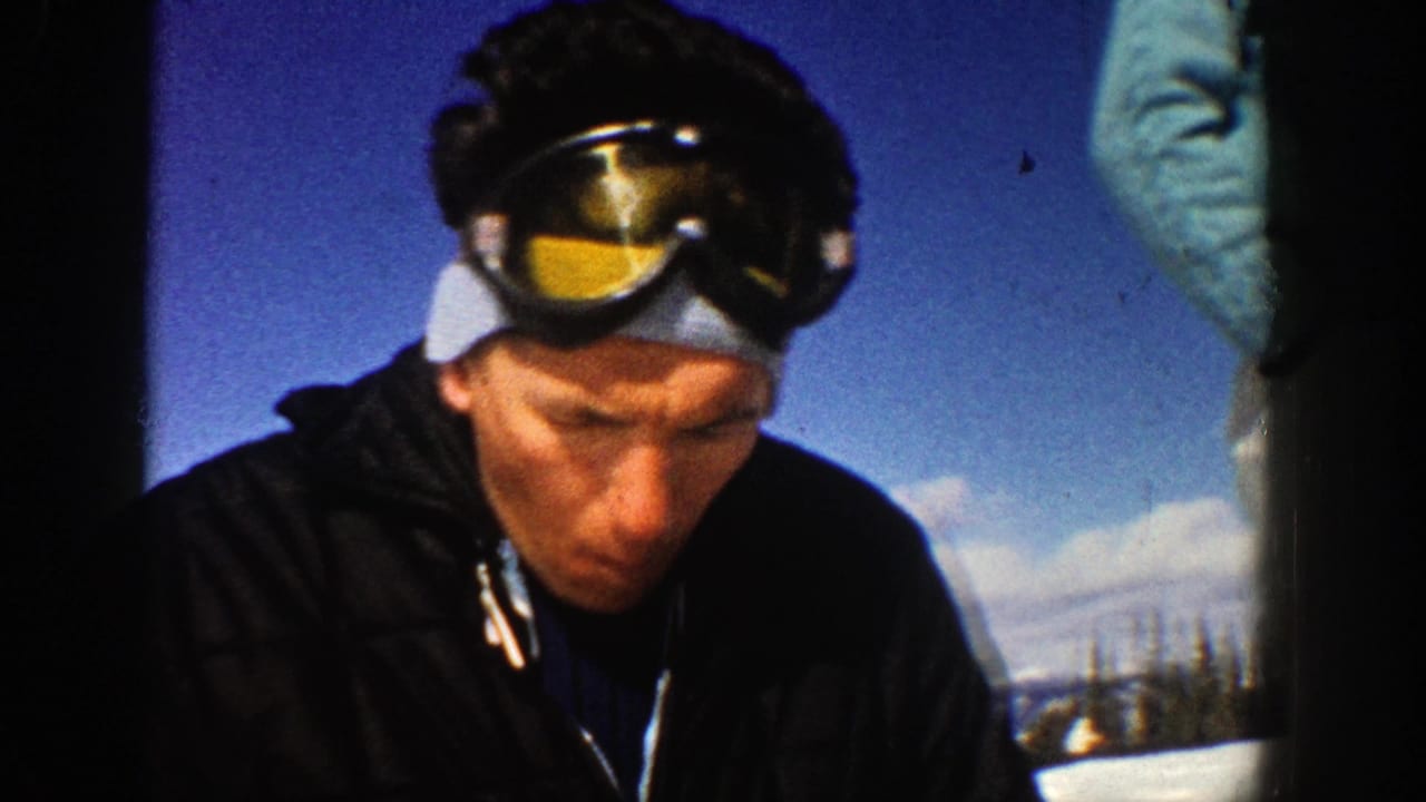 Man in ski goggles and blue cap looks down outdoors against blue sky and snow.
