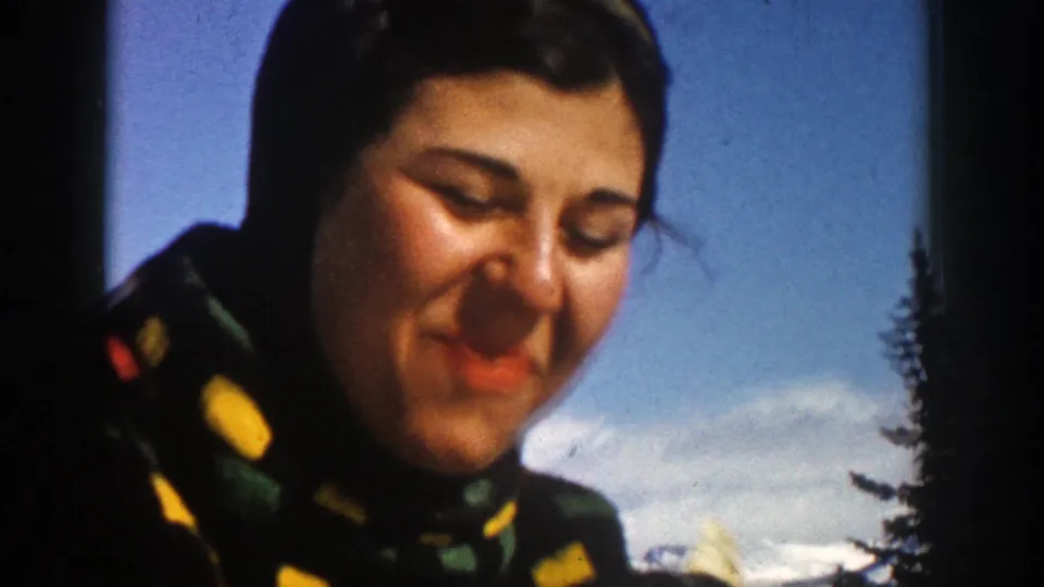1961 Aspen home-movie frame of a smiling woman in a colorful winter jacket against sky and snow.