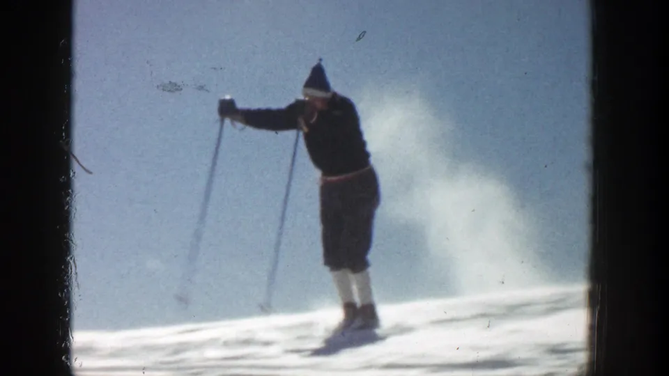 In supported 1961 Aspen footage, a skier in dark clothing and a knit hat moves across a sunlit snowy slope with poles extended.