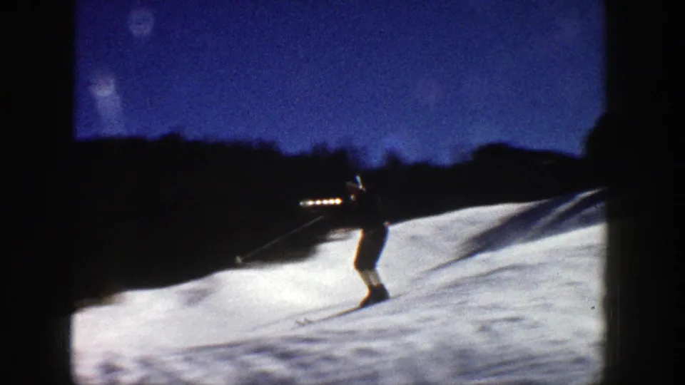 Distant skier on a snow-covered Aspen slope, framed by dark hills and blue sky in 1961 home-movie footage.
