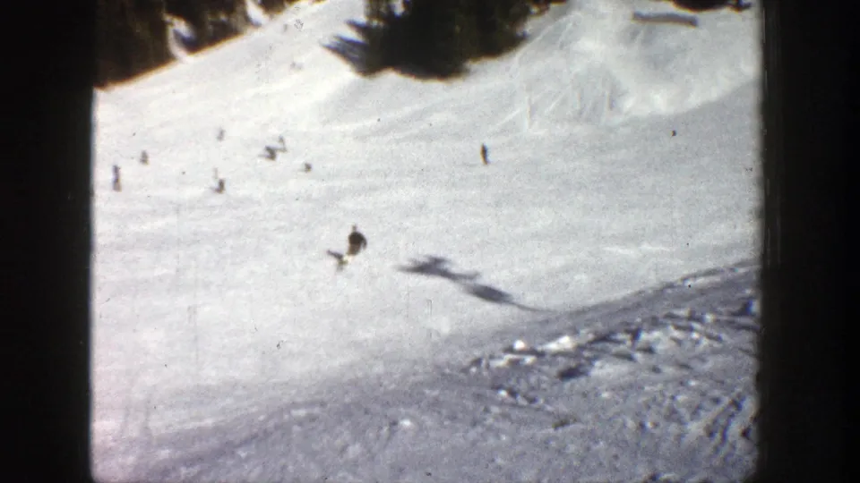 The Aspen 1961 home-movie frame shows multiple skiers spread across a bright slope as one skier crosses the lower area.