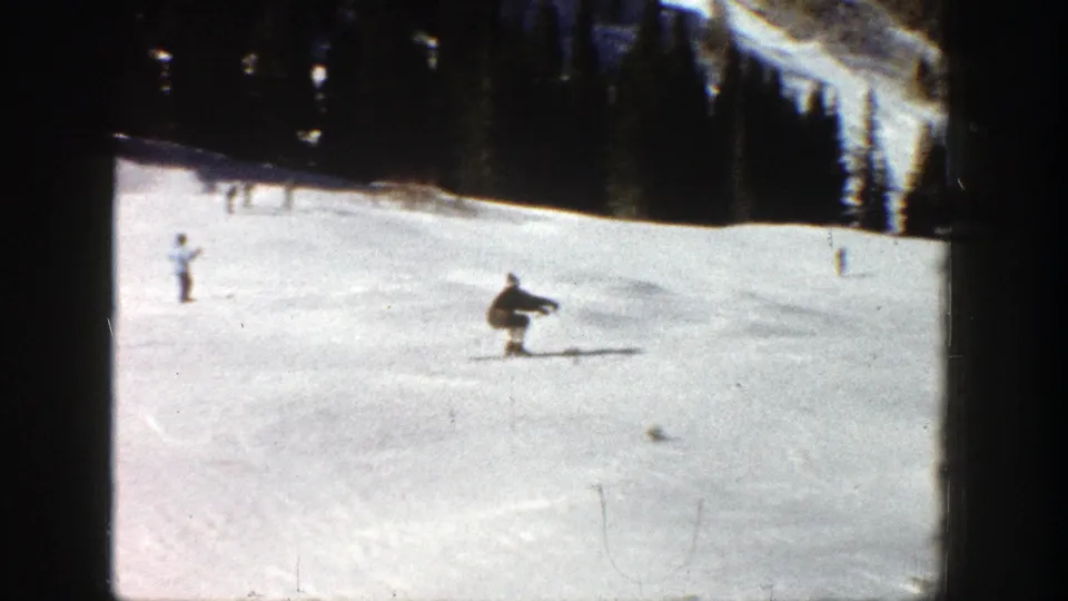 Skier appears crouched during or after a small jump on a 1961 Aspen slope, with dark pines along the edge of the run.