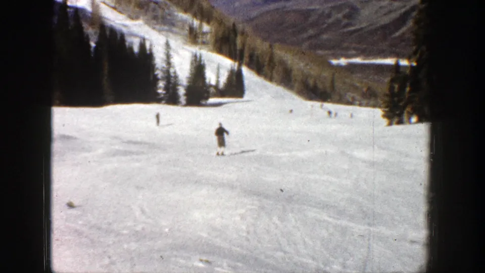 Skier moves away down an Aspen ski run in 1961, framed by pine trees, distant mountain slopes, and 8mm film edges.