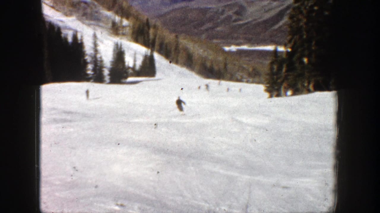 Wide snowy ski slope with several small skiers, evergreen trees, mountains, and dark film borders.