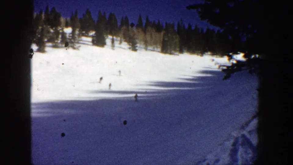 A 1961 Aspen ski slope appears in a wide home-movie view, with scattered skiers below evergreens and visible film dust.