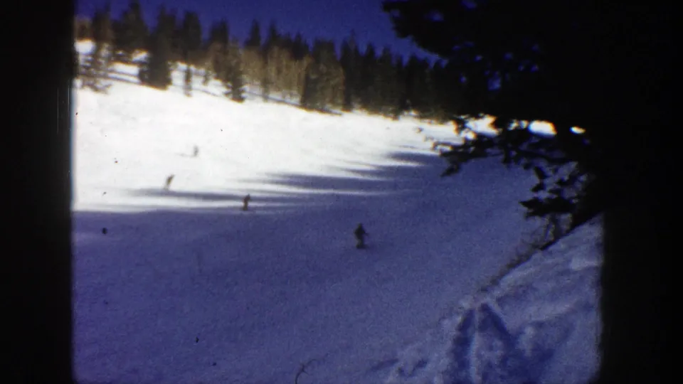 Final 1961 Aspen frame shows a few skiers on a snowy run, with dark foreground branches and evergreen trees along the ridge.