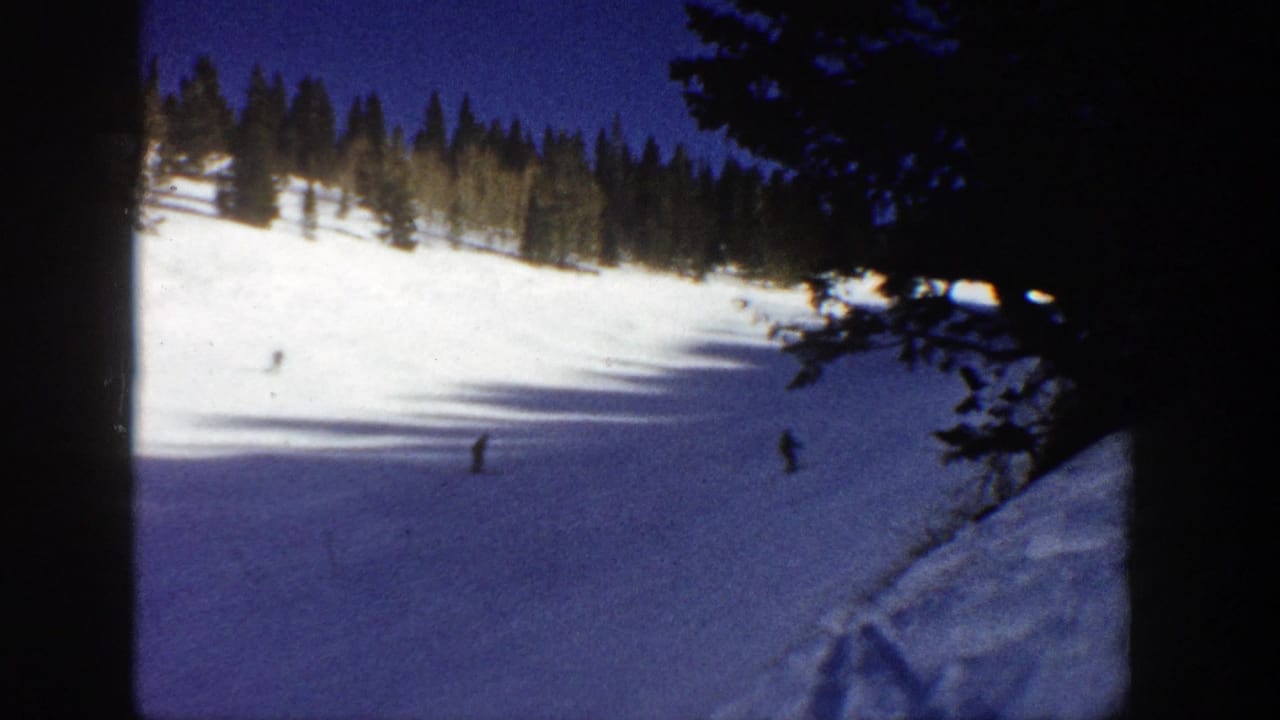 Skiers appear as small figures on a snowy mountainside with evergreens along the upper edge.