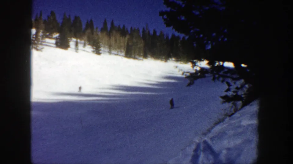 1961 home-movie frame from Aspen showing skiers spaced apart on a bright winter slope, with evergreen trees and dark side framing.