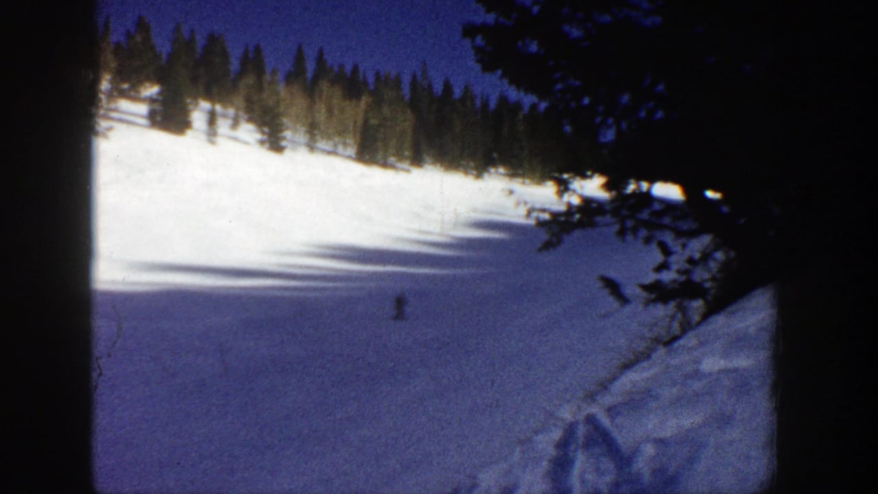 1961 Aspen home-movie frame showing a distant skier crossing a sunlit snow slope with evergreens along the ridge and dark film borders.
